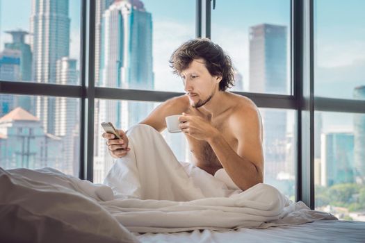 Man wakes up in the morning in an apartment in the downtown area with a view of the skyscrapers and drinks coffee. Life in the noise of the big city concept. Not enough sleep.