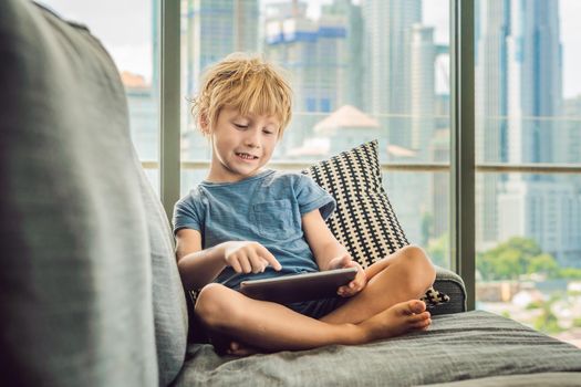 Boy uses a tablet at home on the couch in the background of a window with skyscrapers. Modern children in the megalopolis use a tablet concept.