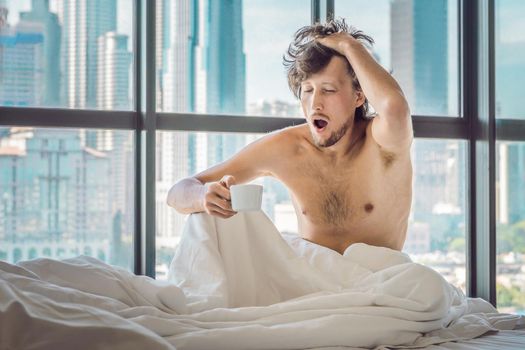 Man wakes up in the morning in an apartment in the downtown area with a view of the skyscrapers and drinks coffee. Life in the noise of the big city concept. Not enough sleep.