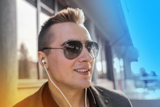Portrait of a positive smiling young businessman of European appearance in sunglasses and headphones listening to music on the street outdoor.