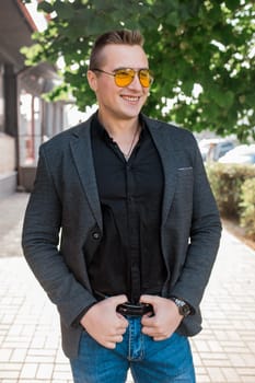 Stylish positive smiling young attractive guy of European appearance businessman portrait in jacket, shirt and jeans, in sunglasses on the street outdoor.