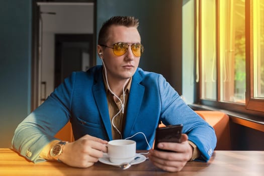 A businessman a stylish portrait of Caucasian appearance in sunglasses, blue jacket, sits at a table on a coffee break in smartphone and headphones, listen to music on cellphone a cafe background.