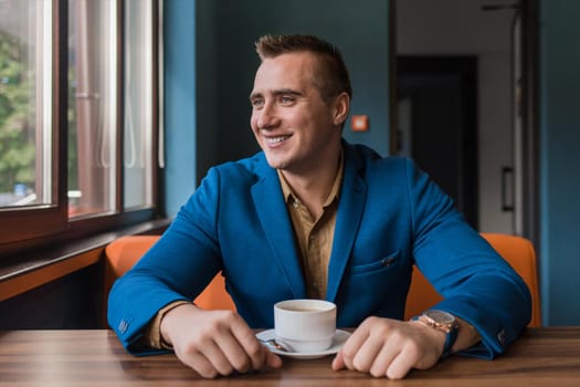 A stylish, positive businessman of Caucasian appearance, a man in a jacket and shirt sits at a table in a cafe on a coffee break and looks away or out the window.