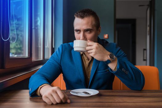 Stylish, adult attractive man of European appearance in a suit sits at a table in a cafe and drinks coffee on a lunch break.