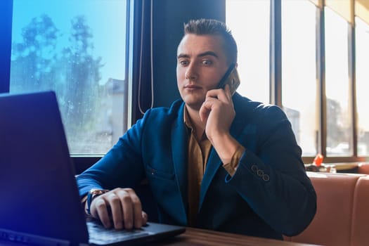 A business man surprised businessman in an attractive European-looking suit works in a laptop, talks on a cell or mobile phone, sitting at a table in a cafe by the window.