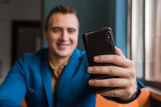 Close-up, businessman guy holds a black smartphone or mobile phone in his hand and takes a selfie or talks on a video link in a cafe.