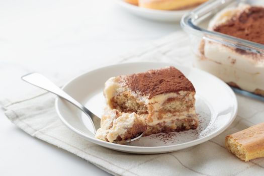 Piece of Homemade Tiramisu cake dessert with savoiardi as ingredients on a white marble background