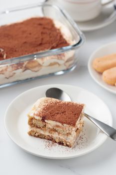 Piece of Homemade Tiramisu cake dessert with savoiardi as ingredients on a white marble background