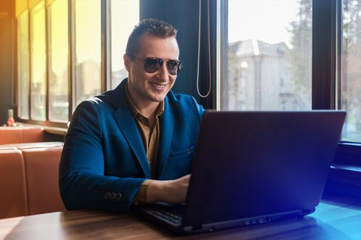 A business man businessman a stylish smiling positive portrait of Caucasian appearance in a blue jacket and sunglasses works in a laptop or computer, sitting at a table by the window in a cafe.