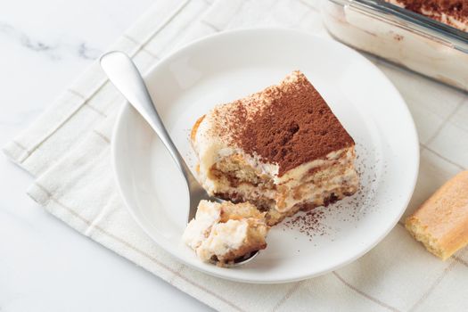 Piece of Homemade Tiramisu cake dessert with savoiardi as ingredients on a white marble background