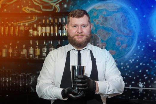 A bearded adult man of European appearance, a professional bartender, holds in his hands in black latex gloves a tool for preparing and mixing alcoholic cocktails in a nightclub.