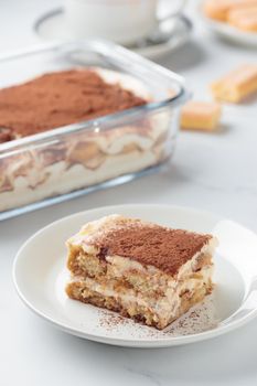 Piece of Homemade Tiramisu cake dessert with savoiardi as ingredients on a white marble background