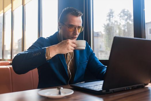 A business man stylish serious businessman in an attractive European-looking suit works in a laptop, listens to music with headphones and drinks coffee sitting at a table in a cafe by the window.