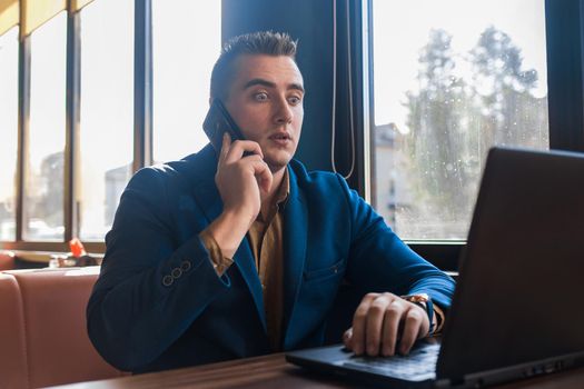 A business man surprised businessman in an attractive European-looking suit works in a laptop, talks on a cell or mobile phone, sitting at a table in a cafe by the window.