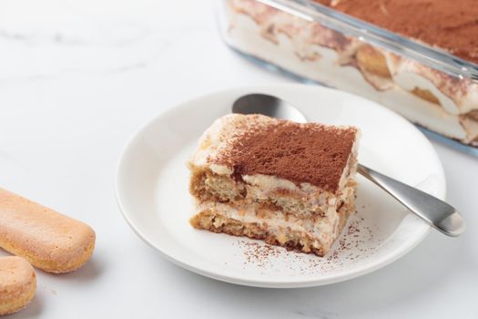 Piece of Homemade Tiramisu cake dessert with savoiardi as ingredients on a white marble background