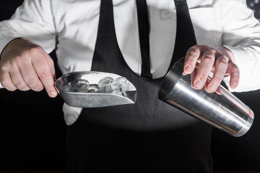 The hands of a professional bartender add ice in a scaple to a tool for preparing and mixing alcoholic cocktails, a metal shaker.