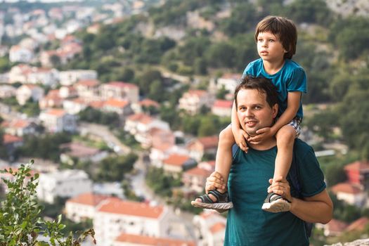 Father holding his little son on the sholders. Family hiking in the mountains with the panoramic view of the old Mediterranean city behind them. Kotor Bay in Montenegro