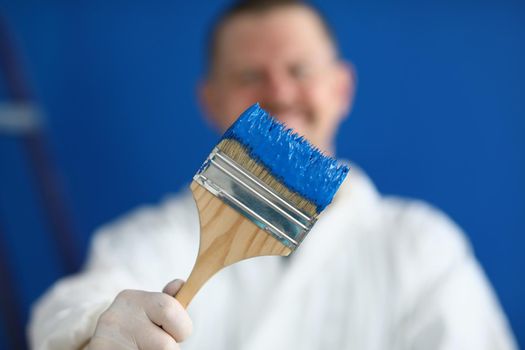 Close-up of painter worker hold brush covered in blue colour paint, handyman on construction site. Renovation, design, construction site, project concept