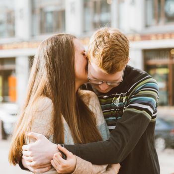 Portrait of happy couple embracing in downtown, red-haired man with glasses kisses and woman with long hair. Girl whispers in the ear of the guy. Concept of teenage love and first kiss, love, relationship, couple. City, waterfront.