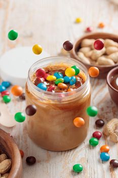 Jar with peanut butter and sweet ingredients on white wooden background closeup