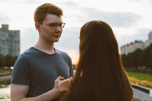 Girl with long thick dark hair holding hands redhead boy in blue t-shirt on bridge, teen love at evening. Boy looks tenderly at girl, young couple. Concept of a teenage love and first kiss, sincere feelings of man and woman, city, waterfront. Close up