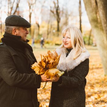 Happy blonde mature woman and handsome middle-aged brunette man walk in park, looking at each other. Loving couple of 45-50 years old walks in the autumn park in warm clothes, holding bouquet of leaves