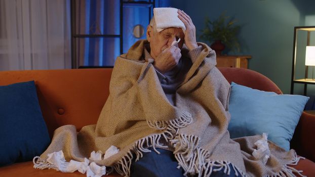 Portrait of one senior grandfather man suffering from cold or allergy in plaid sitting on sofa in living room at home. Sick elderly grandpa with towel on head blows nose snot into napkin. Coronavirus