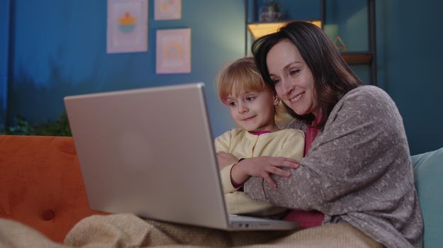 Happy parent mother and cute small child daughter kid looking at laptop screen having fun watching funny study movies or learn together at home relaxing laughing lying on sofa. Family of two people