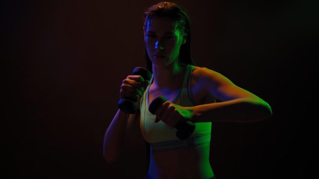 Sportive girl posing doing hand workout biceps with dumbbells standing in neon lights wearing sport top working on a arm weight hit isolated on black background. Neon light concept.