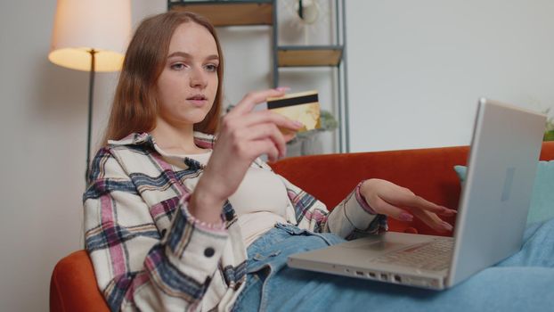 Smiling Caucasian young woman lying on sofa using credit bank card and laptop computer while transferring money, purchases online shopping in living room home. Female girl tapping, scrolling, browsing