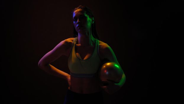 Professional athlete girl posing with power exercise ball standing in neon lights wearing sport top working on a weight fitness isolated on black background. Neon light concept.