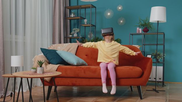 Exited Caucasian amazed toddler girl sitting on sofa in living room using headset helmet app to play simulation game. Watching virtual reality 3D 360 video. Teen school kid at home alone in VR goggles