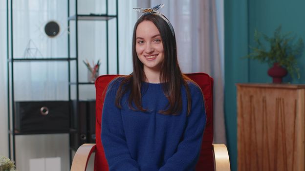 Portrait of happy one beautiful teenager student girl in blue sweater smiling, looking at camera. Young cute fashion model woman indoors isolated at home in living room on chair. Female nature beauty