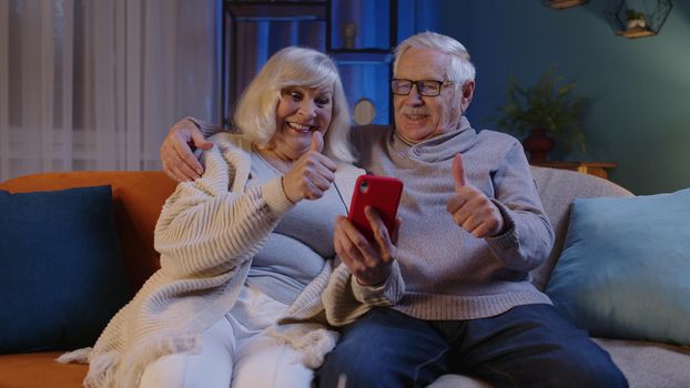 Happy senior elderly couple grandfather and grandmother looking on mobile phone making online shopping and smiling. Mature family husband wife watching news videos, using social media at home