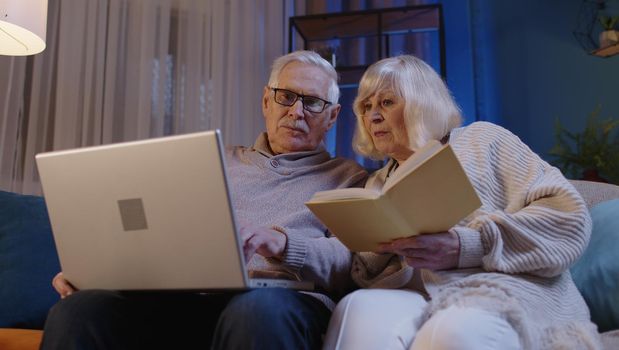 Happy senior grandparents couple relaxing enjoying leisure hobbies on couch in living room at home, elderly wife grandma reading book while older husband grandpa using laptop pc receiving bad news