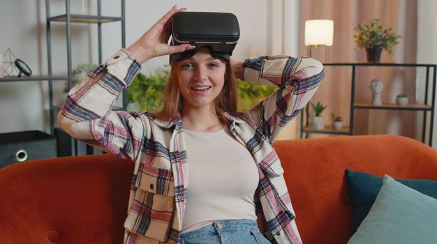 Young woman use VR headset glasses at home, enjoying 3D 360 video concept, entertaining, in positive mood, moving hands in air, sitting on sofa in living room. Virtual reality, modern gadget device