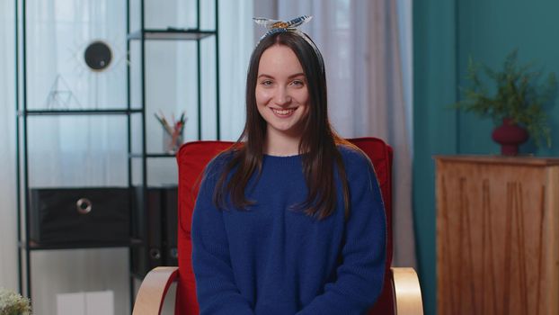 Portrait of happy one beautiful teenager student girl in blue sweater smiling, looking at camera. Young cute fashion model woman indoors isolated at home in living room on chair. Female nature beauty
