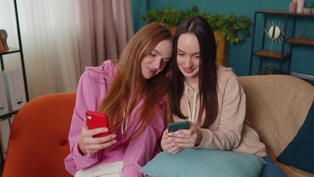 Excited girls friends siblings watching photos on two smartphones, online dating app. Happy female women couple family on sofa at flat home, enjoy chatting, spend leisure time on social media network