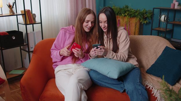 Cheerful girls friends siblings watching photos on two smartphone, online dating app. Happy female women couple family on sofa at flat home, enjoy chatting, spend leisure time on social media network