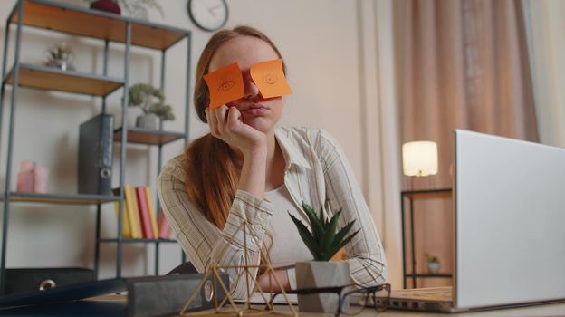 Tired woman freelancer using laptop computer napping at home office desk with funny stickers on eyes sleeps at workplace, suffering migaine headache after e-learning, remote working. Employee burnout