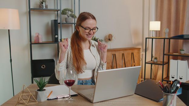 Young woman freelancer gets good news, positive exam results, new opportunity, successfully signed contract, celebrates victory drinking wine after working on laptop computer sitting at home office