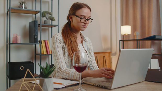 Young woman freelancer drinking wine from glass while working typing on laptop computer sitting at desk at home office. Focused female girl businesswoman working from distant workplace, remote job