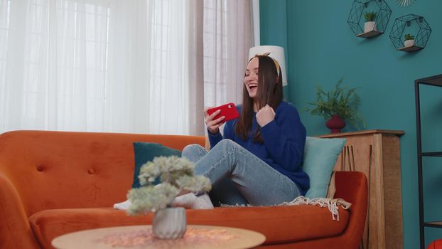 Worried funny addicted woman enthusiastically playing racing video games on mobile phone at home in living room on sofa. Young adult teen girl using smartphone gadget app with drive simulator isolated