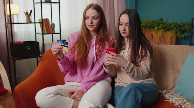 Cheerful girls friends siblings using credit bank card and smartphone while transferring money, purchases online shopping. Happy two female women couple family on sofa at home, spending time together