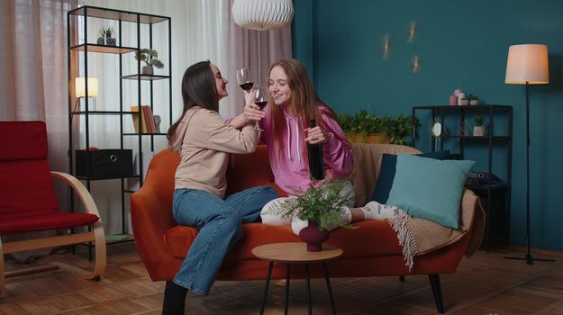 Joyful girls friends siblings clinking glasses of red wine cheering and drinking, celebrate birthday party hangout at home in living room. Two female women couple family celebrating new house purchase