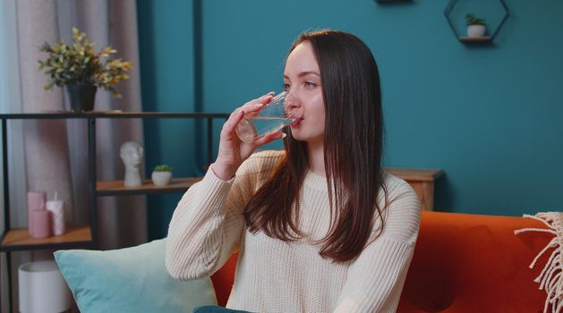 Close-up portrait of young woman sitting on sofa at home holds glass drinks water, makes gulp of clean aqua reducing thirst caring about health and beauty. Healthy girl lifestyle, enough liquid intake