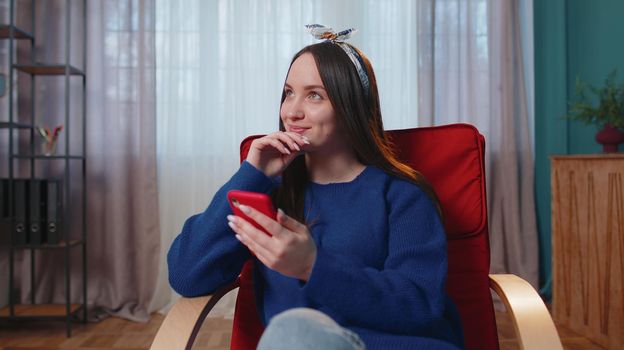Cheerful relaxing young brunette woman sitting on orange chair uses mobile phone smile at modern home apartment. Happy girl texting share messages content on smartphone social media application online