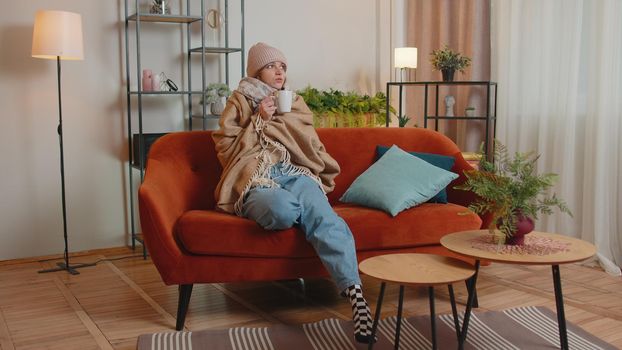 Young sick woman wear hat wrapped in plaid sit alone shivering from cold on sofa drinking hot tea in unheated apartment without heating due debt. Unhealthy female feeling discomfort try to warming up