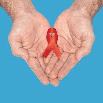 Red awareness ribbon bow on mans helping hands isolated on blue background. HIV, AIDS world day. Social life issues concept. Aids charity fund concept. Healthcare and medicine concept.