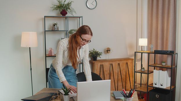 Young professional Caucasian woman in shirt, standing by desk in home office, looking at paper documents analyzes checks data, calculating incomes outcomes working on laptop computer. Distance job